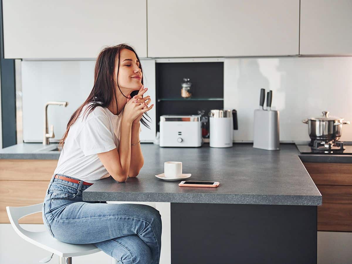 Uma jovem sentada na sua moderna cozinha tipo península, a saborear um café com os olhos fechados. O mobiliário é branco com pormenores em madeira e a bancada é cinzenta.