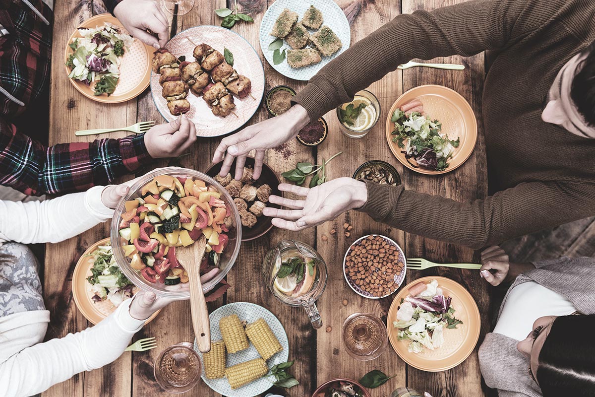 Vista aérea de uma reunião de mesa de jantar onde são partilhados diferentes pratos como milho, salada, espetadas de carne, etc.