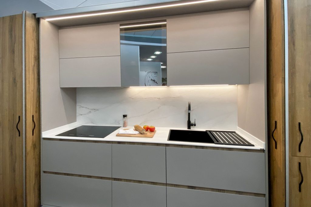 Vista frontal de uma pequena cozinha branca com placa vitrocerâmica integrada e lava-loiça preto, espelho entre os móveis de parede e luz LED por baixo.