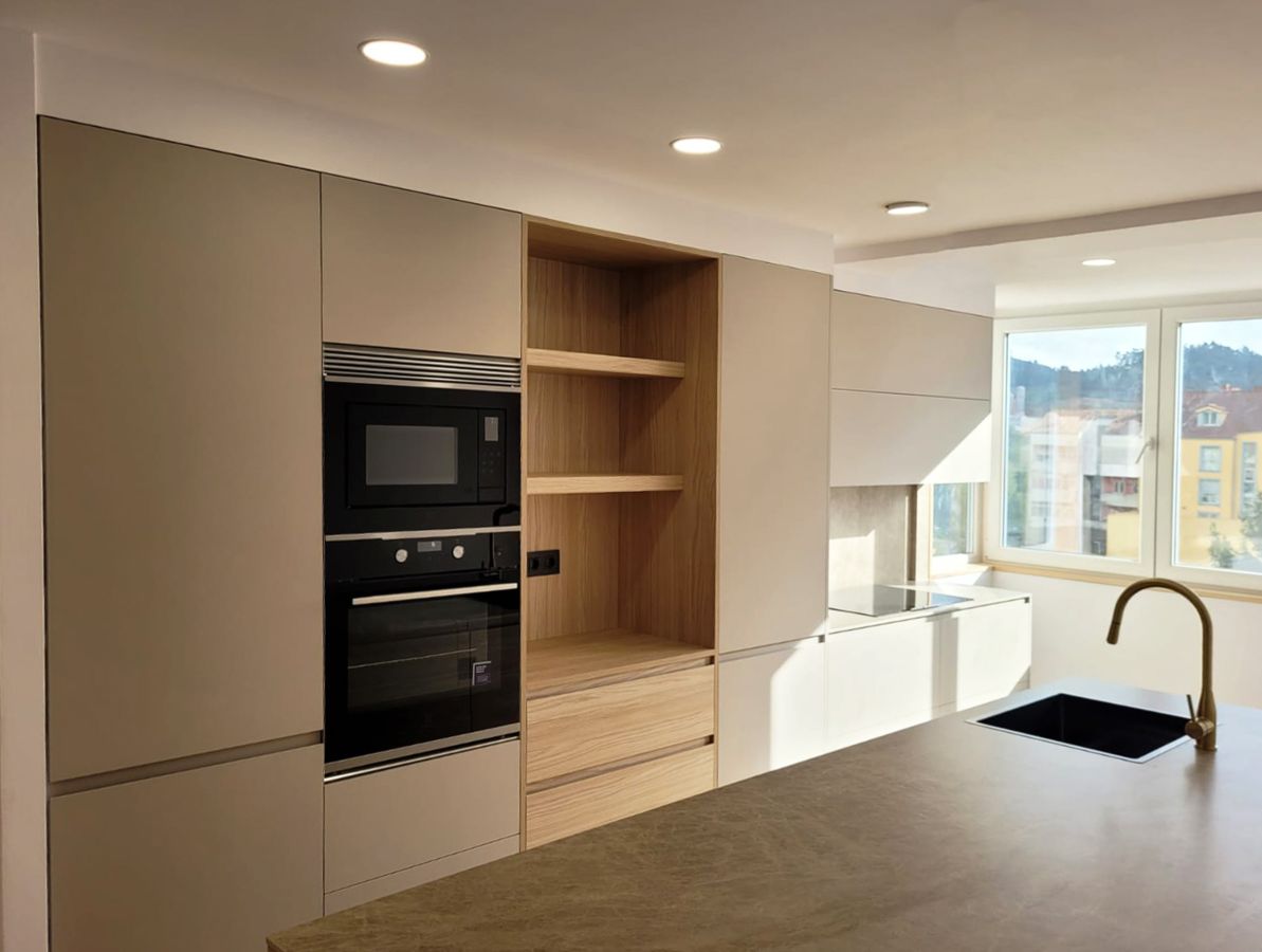 Cozinha cinzenta iluminada equipada com forno, micro-ondas e outros eletrodomésticos integrados e com um armário de madeira aberto no centro.