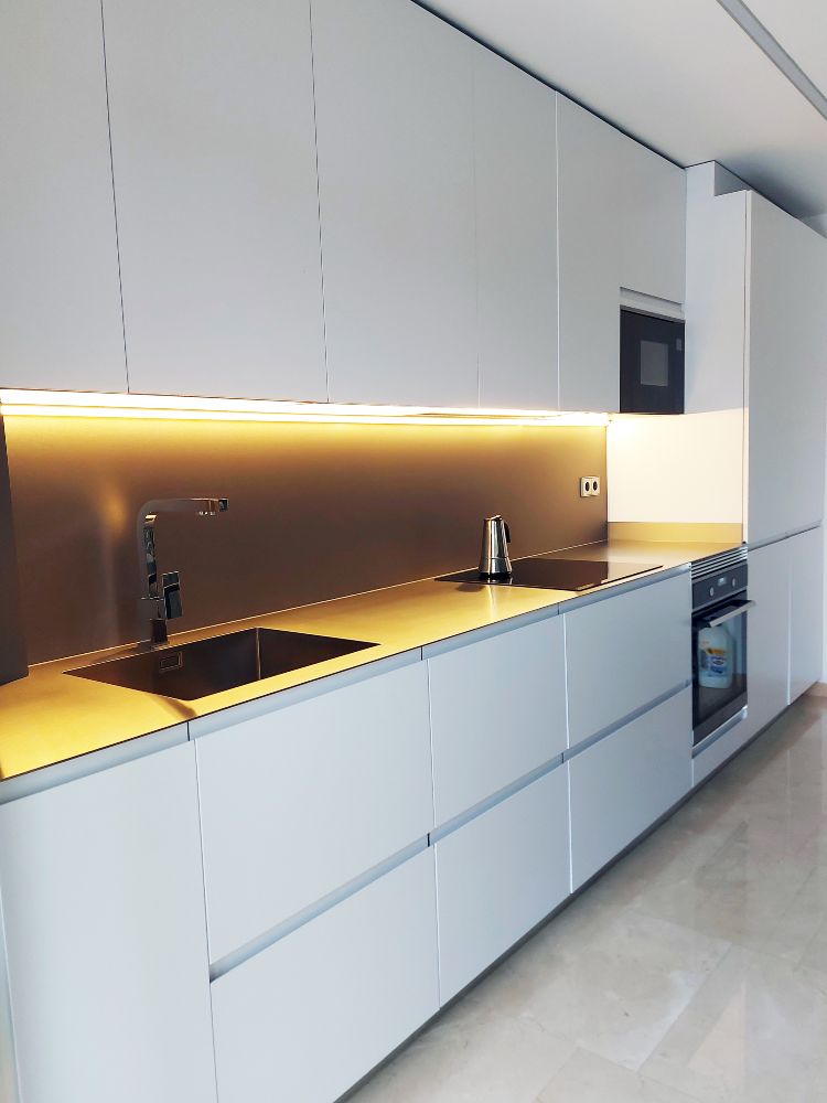 Vista lateral da frente da cozinha branca com forno e micro-ondas integrados, placa vitrocerâmica e lava-loiça e iluminação LED na base dos móveis de parede.