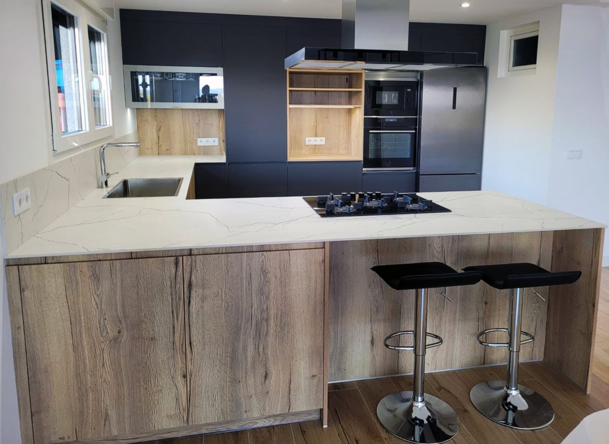 Vista frontal da cozinha em forma de U com móveis em madeira e cor preta, com placa, exaustor, lava-loiça e electrodomésticos integrados.