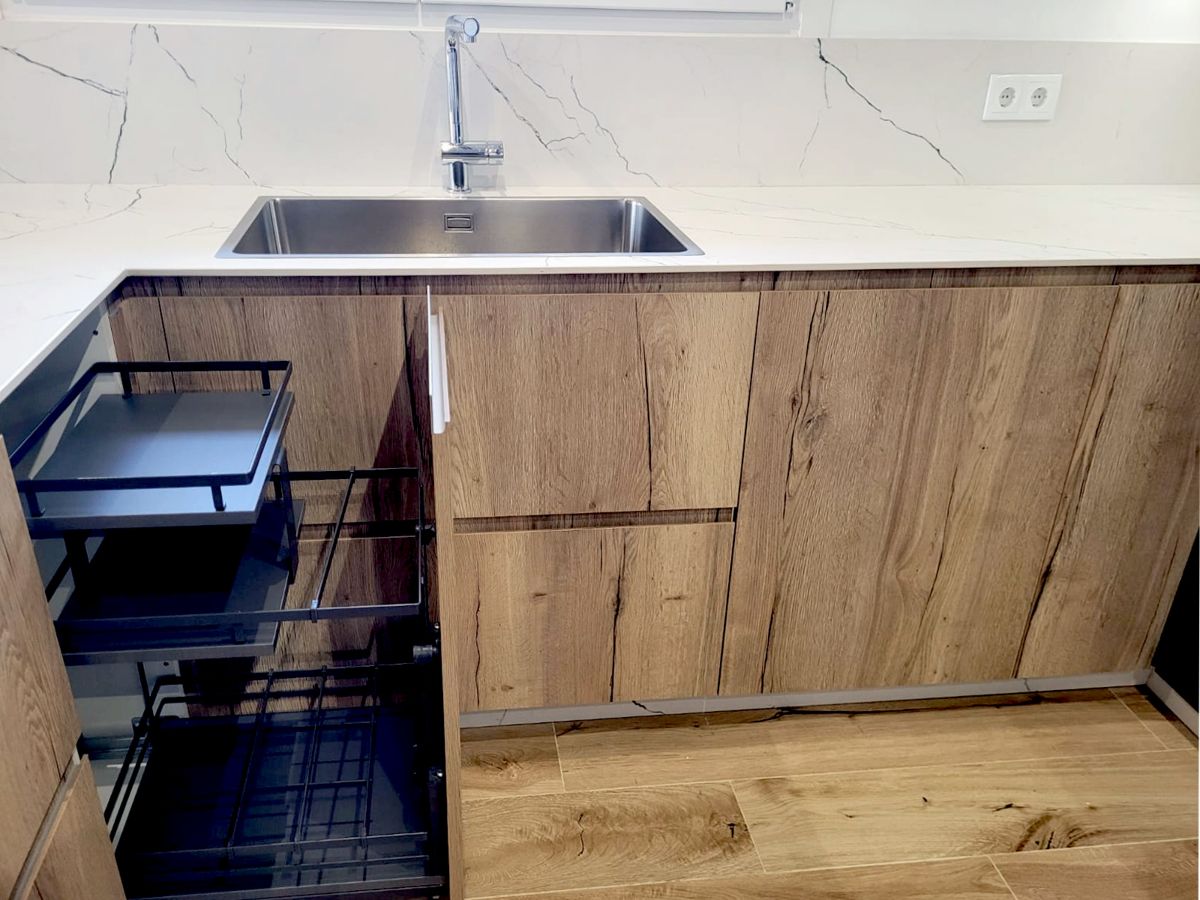 Pormenor da bancada de cozinha em porcelana branca com lava-loiça integrado e torneira prateada, com móveis inferiores em madeira clara, um deles aberto.