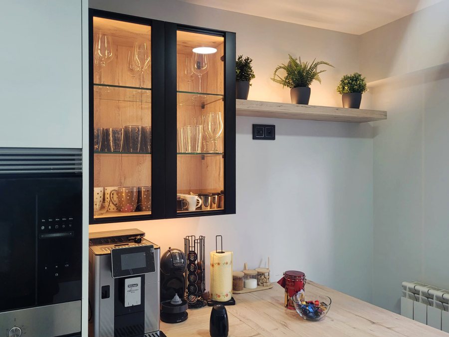 Cozinha moderna a preto e branco com armário de vidro com portas pretas e prateleiras de vidro com chávenas e copos organizados no interior.