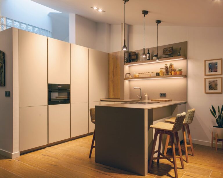 Cocina abierta al salón con península central, muebles en Beige Oliva modelo Fussion de Senssia, encimera Silestone y estanterías iluminadas con acabado en roble Rakia.